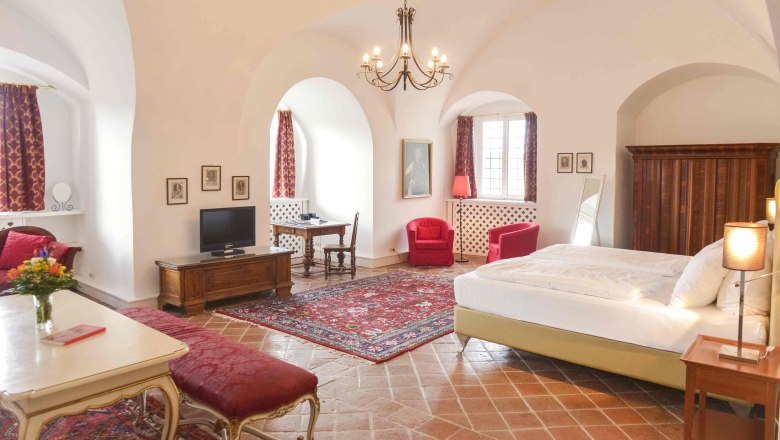 Luxussuite, © Schlosshotel Mailberg / point of view Luxuriöse Suite mit antiken Möbeln, roten Teppichen und Vorhängen, einem großen Bett und einem Kronleuchter.