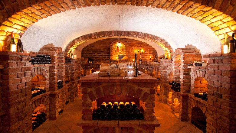 Weinkeller - Vinothek, © Familie Pichler Ein stimmungsvoll beleuchteter Weinkeller mit Backsteinwänden und Weinflaschen in Regalen.