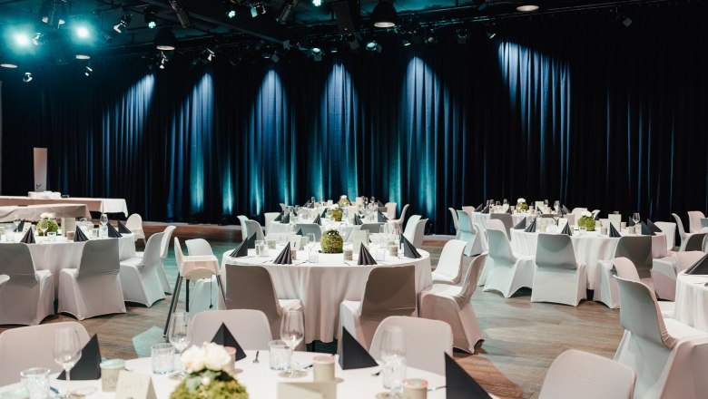 MOMENT, Galabestuhlung, © diebelichterei.at Elegante Galabestuhlung mit runden Tischen, weißen Tischdecken und schwarzen Servietten in einem festlich beleuchteten Saal.