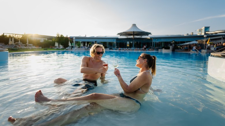 Therme Laa, Außenbereich, © Thermenresort Laa / Pflanzl Media Zwei Personen entspannen im Pool des Thermenresorts Laa und stoßen mit Weingläsern an.