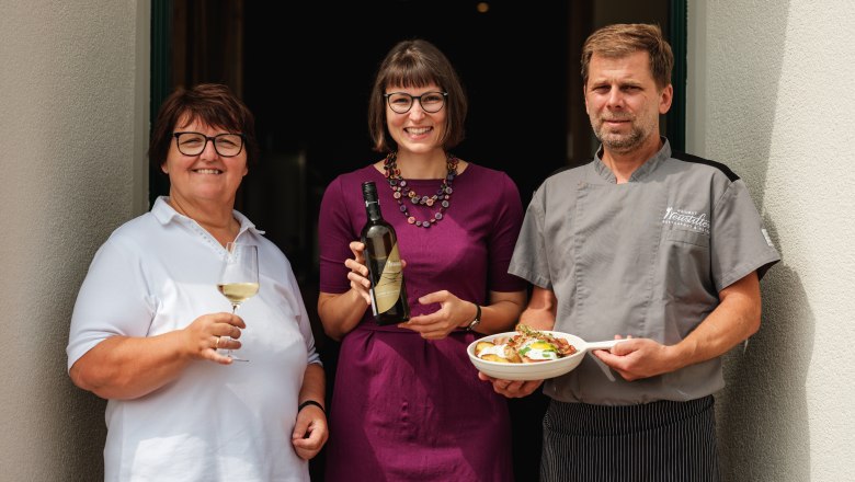 Wein.Restaurant Neustifter, © Weinviertel Tourismus / Michael Reidinger Drei Personen vor einem Restaurant, eine Frau mit Weinglas, eine Frau mit Weinflasche, ein Mann im Kochoutfit mit Teller.