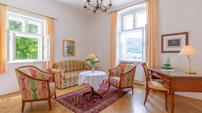 Hotelsuite im Hotel Richard Läwenherz, © Chris Saupper Elegantes Wohnzimmer mit antiken Möbeln, zwei Fenstern, einem Tisch mit Blumen, einem Schreibtisch und einem Teppich auf Holzboden.