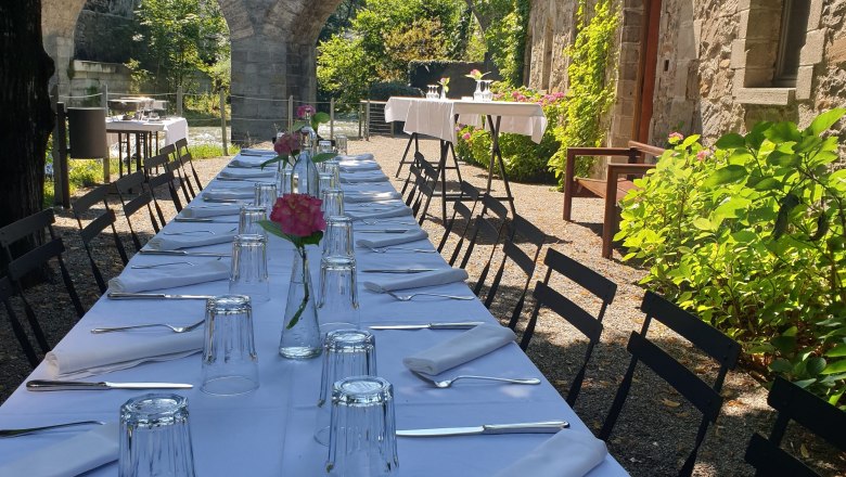 Schloss Rothschild, Schwarze Kuchl, © Waidhofen an der Ybbs Langer Tisch im Freien mit weißer Tischdecke, gedeckt mit Gläsern und Besteck, im Hintergrund Steinbögen und grüne Vegetation.