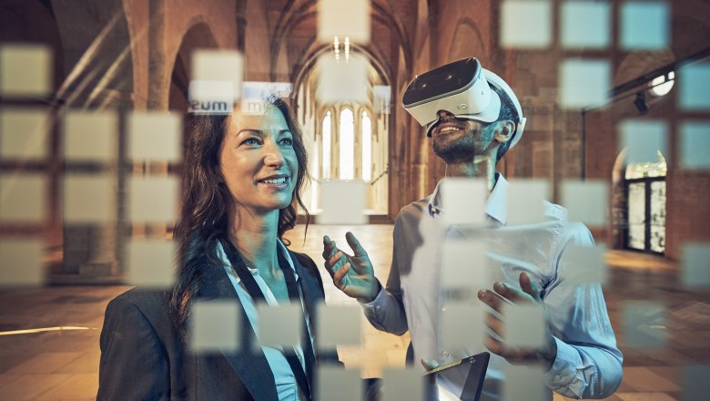 Convention Partner Krems, © Andreas Hofer Zwei Personen in einem historischen Gebäude, eine trägt eine VR-Brille, die andere lächelt. Im Vordergrund sind unscharfe Quadrate zu sehen.