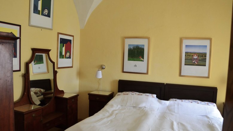 Ferienwohnung Rossarium Schlafzimmer 2, © Renaissanceschloss Greillenstein Schlafzimmer mit Doppelbett, gelben Wänden und mehreren gerahmten Bildern an der Wand.