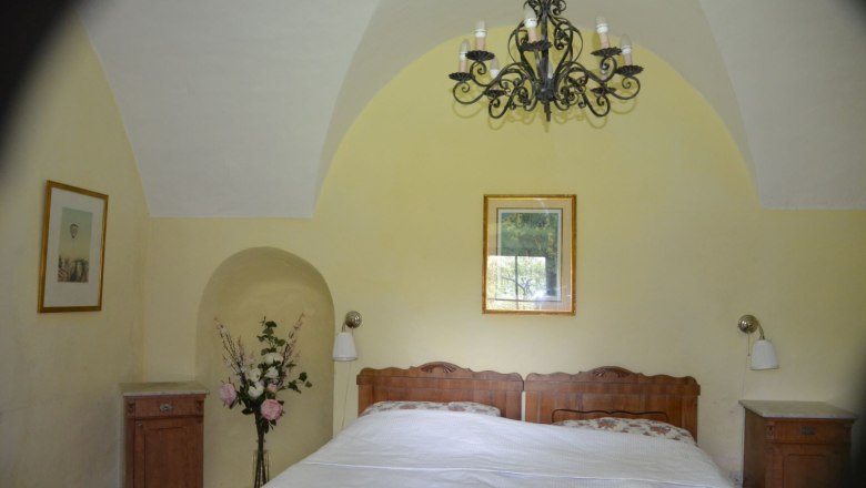 Ferienwohnung Rossarium Schlafzimmer 1, © Renaissanceschloss Greillenstein Schlafzimmer mit Doppelbett, gelben Wänden, einem Kronleuchter und Blumen in einer Vase.