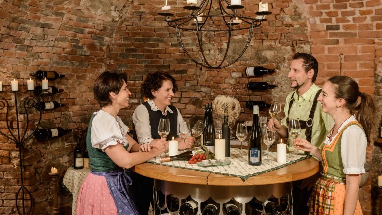 Weinkeller im Hotel-Restaurant Zur Post, © BOW Rita Newman Vier Personen in traditioneller Kleidung stehen in einem Weinkeller und halten Weingläser. Sie lachen und unterhalten sich an einem runden Tisch.