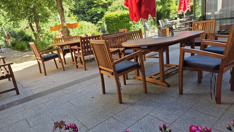 Flackl-Wirt - Gastgarten, © Flackl - Wirt GmbH & Co KG Ein gemütlicher Gastgarten mit Holzmöbeln und roten Sonnenschirmen, umgeben von grünen Bäumen und Blumen.