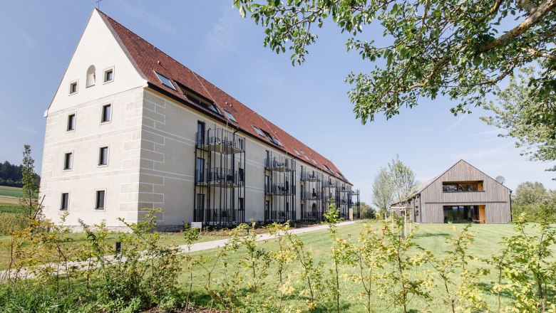 Korngut, © Holter Ein großes, modernes Gebäude mit rotem Dach und Balkonen neben einem kleineren Holzhaus auf einem grünen Grundstück.