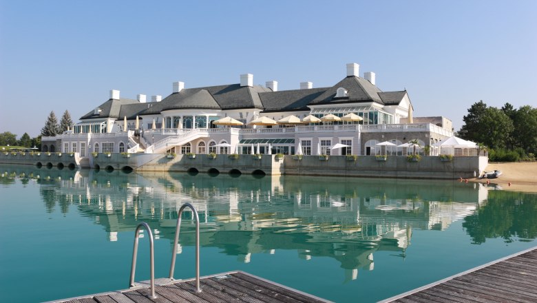 Fontana Golfclub, Clubhaus, © Fontana GmbH Ein elegantes Clubhaus am See mit Terrasse und Sonnenschirmen, umgeben von klarem Wasser und einem Steg im Vordergrund.