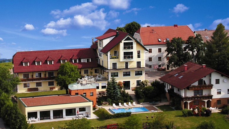 Hotel des Glücks, © Hotel des Glücks, Hermann Cisar Ein Hotelkomplex mit roten Dächern, Pool und Garten, umgeben von Bäumen und blühenden Pflanzen unter einem blauen Himmel.
