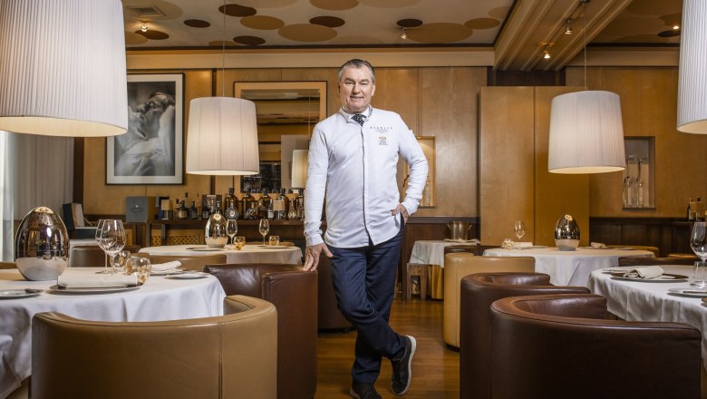 Die Welt von MÖRWALD, Toni Mörwald, © Lukas Aigelsreither Ein Mann in weißer Kochjacke steht in einem eleganten Restaurant mit runden Tischen und großen Lampen.