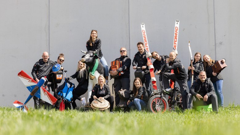 RETTER EVENTS, Team, © RETTER EVENTS Gruppe von Menschen posiert mit Sportgeräten und einer Kuhstatue auf einer Wiese.