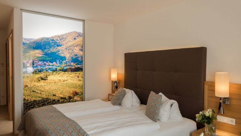 Donauhof Doppelzimmer, © Marcella Wallner Modernes Hotelzimmer mit Doppelbett, dekorativen Kissen und großem Landschaftsbild an der Wand.