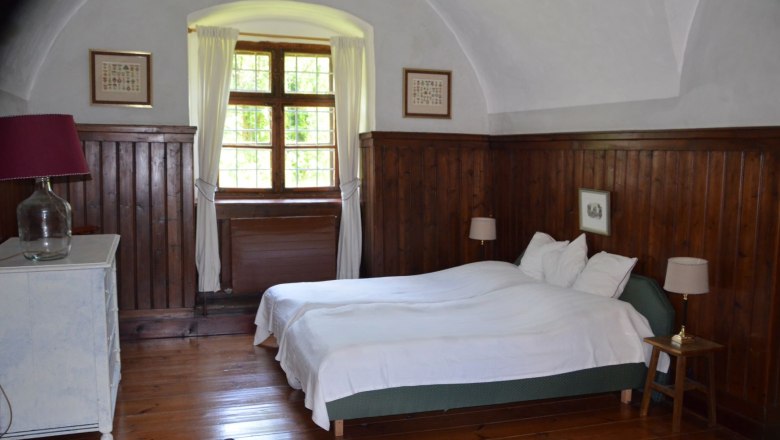 Ferienwohnung Vogtei Schlafzimmer, © Renaissanceschloss Greillenstein Schlafzimmer mit Holzwänden, einem Doppelbett, einem Fenster mit Vorhängen und einer Kommode mit Lampe.