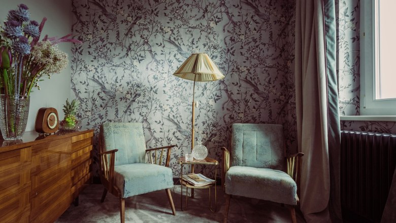 Zimmer, © Matthias Kronfuss Ein stilvoll eingerichtetes Zimmer mit zwei blauen Sesseln, einer Stehlampe und einer Kommode mit Blumen und einer Uhr.