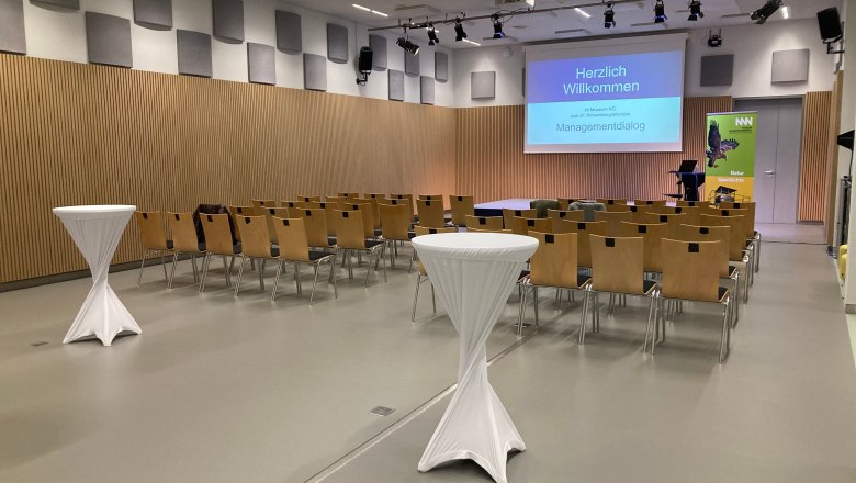 Museum Niederösterreich, Museumslounge, © NÖ Museum Betriebs GmbH, Foto: Heike Brantner Konferenzraum mit Stuhlreihen, Stehtischen und einer Leinwand mit der Aufschrift 'Herzlich Willkommen'.