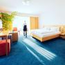 Wohnraum_3, © Franz Klinglhuber GmbH Helles Hotelzimmer mit Doppelbett, Schreibtisch, Fernseher und großer Pflanze. Eine Person geht durch den Raum.