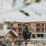 Naturhotel Molzbachhof, © Heldentheater, Hotel Molzbachhof Ein modernes Holzgebäude mit Balkonen und ein traditionelles Holzhaus im Vordergrund, umgeben von Bäumen und Gartenanlagen.