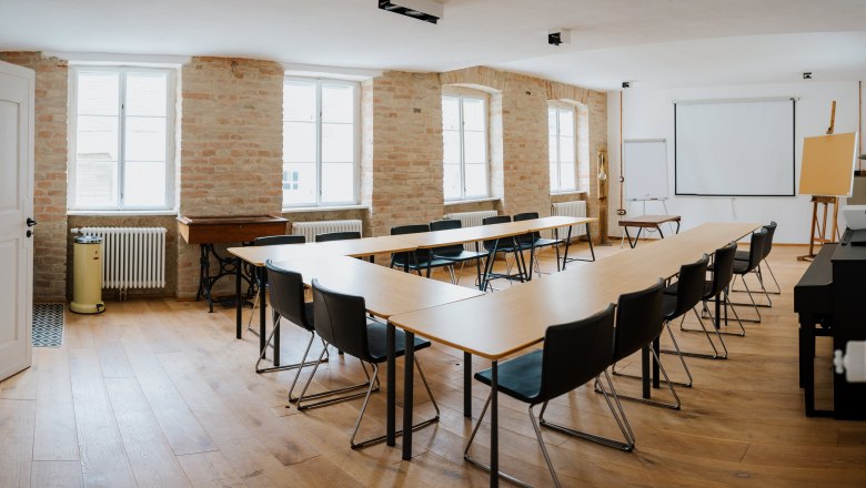 Business Forge, © Voglperspektive Ein heller Seminarraum mit U-förmiger Tischanordnung, Stühlen, Projektionsleinwand und Flipchart.