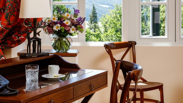 Zimmer Knappenhof, © Knappenhof - Christian Husar Ein elegantes Zimmer mit Holzmöbeln, einem Schreibtisch, Stühlen und einem Fenster mit Blick auf eine grüne Landschaft.