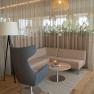 Moderne Sitzecke mit grauem Sessel, gemustertem Sofa, rundem Holztisch und Stehlampe vor einem Vorhang.