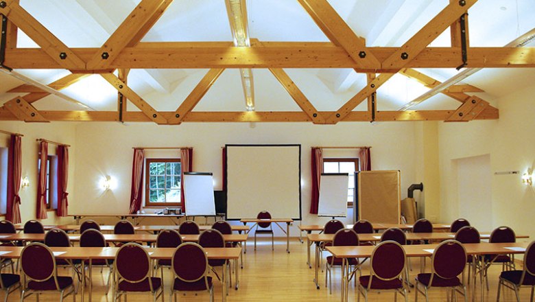 Seminarraum mit Holzbalkendecke, Tischen und Stühlen, Leinwand und Flipcharts.
