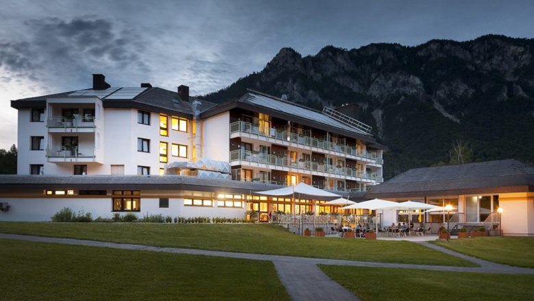Ein beleuchtetes Hotelgeb&auml;ude vor einer Bergkulisse in der Abendd&auml;mmerung.