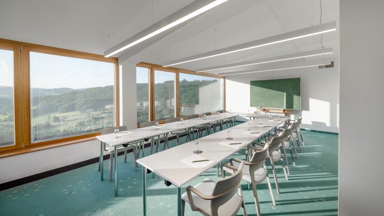 Ein heller Seminarraum mit gro&szlig;en Fenstern, die eine Aussicht auf eine gr&uuml;ne Landschaft bieten. Wei&szlig;e Tische und St&uuml;hle sind in U-Form angeordnet.