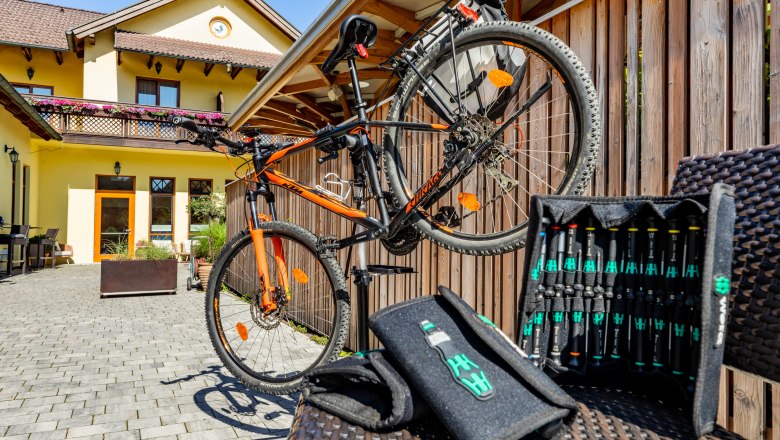 Ein orangenes Fahrrad auf einem St&auml;nder vor einem gelben Geb&auml;ude mit Holzzaun und Werkzeugset im Vordergrund.