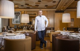 Ein Mann in weißer Kochjacke steht in einem eleganten Restaurant mit runden Tischen und großen Lampen.