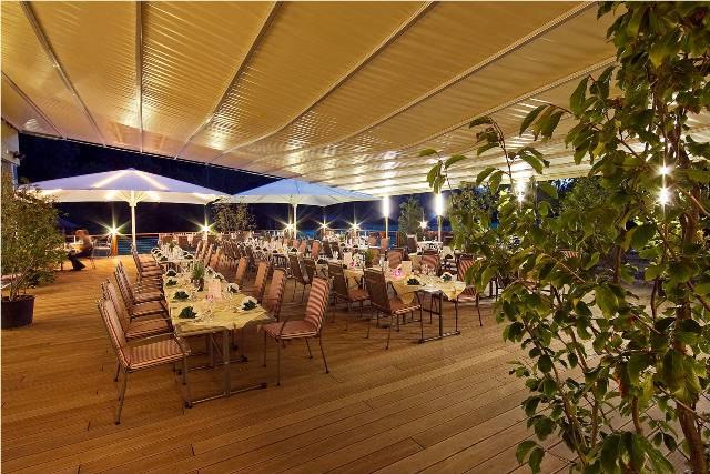 Eine festlich gedeckte Terrasse mit Tischen und Stühlen unter einem großen Sonnenschutz bei Nacht.