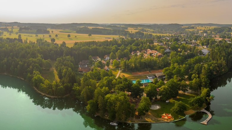 Luftaufnahme eines Sees mit umliegendem Wald und Geb&auml;uden.