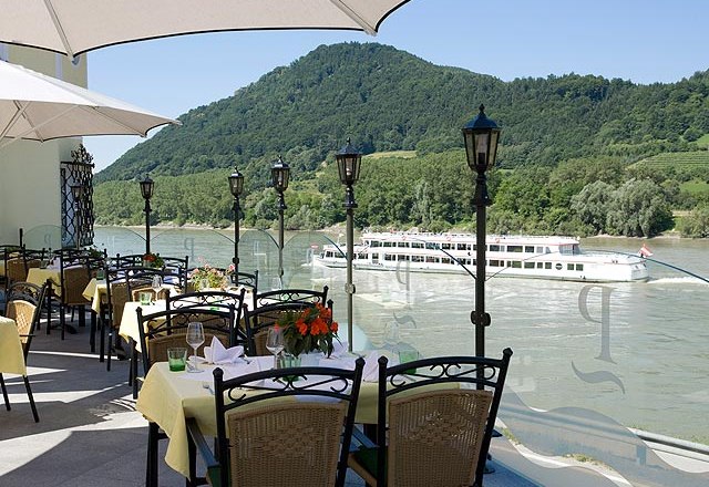 Terrasse mit gedeckten Tischen, Blick auf Fluss und vorbeifahrendes Schiff, umgeben von gr&uuml;nen H&uuml;geln.
