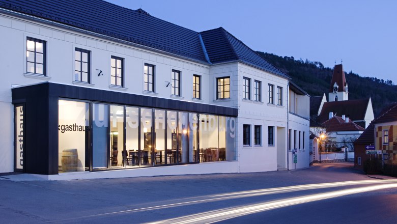 Au&szlig;enansicht des Gasthofs Hotel Zur Schonenburg bei D&auml;mmerung mit beleuchteten Fenstern und vorbeifahrenden Autos.