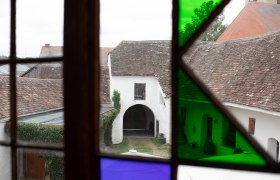 Blick durch ein Fenster mit buntem Glas auf einen alten Innenhof mit gewölbtem Tor und Ziegeldächern.