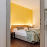 Hotelzimmer mit gelber Wand, Doppelbett und Pflanze im Vordergrund.