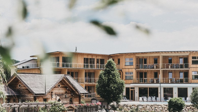 Ein modernes Holzgeb&auml;ude mit Balkonen und ein traditionelles Holzhaus im Vordergrund, umgeben von B&auml;umen und Gartenanlagen.