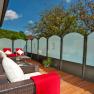 Eine Terrasse mit Rattanm&ouml;beln, roten Decken und Kissen, umgeben von Glasw&auml;nden und B&auml;umen im Hintergrund.