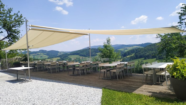 Terrasse mit Sonnensegel und Tischen, umgeben von grüner Landschaft und blauem Himmel.