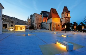Abendaufnahme der beleuchteten Burg Perchtoldsdorf mit modernem Vorplatz und Sitzbänken.