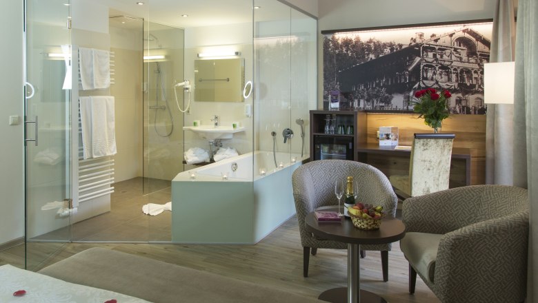Luxuriöses Hotelzimmer mit Glasbad, gemütlichen Sesseln, Tisch mit Champagner und Früchten, sowie einem großen Bett mit Rosenblättern.
