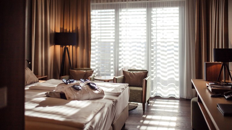 Hotelzimmer mit Doppelbett, Badem&auml;nteln, zwei Sesseln und einem Tisch vor einem gro&szlig;en Fenster mit Vorh&auml;ngen.