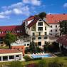 Ein Hotelkomplex mit roten D&auml;chern, Pool und Garten, umgeben von B&auml;umen und bl&uuml;henden Pflanzen unter einem blauen Himmel.