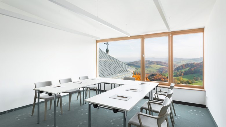 Ein heller Seminarraum mit weißen Tischen und Stühlen, großen Fenstern mit Blick auf eine hügelige Landschaft.