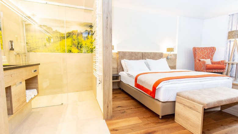 Modernes Hotelzimmer mit Doppelbett, orangefarbenem Sessel und offener Dusche.