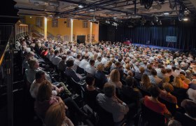Ein gefüllter Theatersaal mit Publikum, das auf eine Bühne blickt.