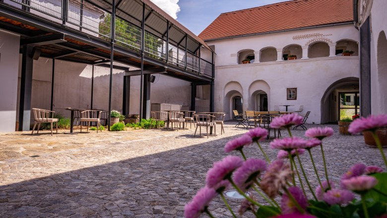 Ein Innenhof mit Kopfsteinpflaster, Tischen und St&uuml;hlen, umgeben von Geb&auml;uden mit Arkaden und einem modernen Balkon.