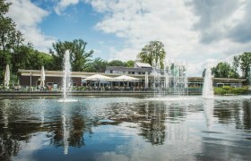 Ein modernes Geb&auml;ude mit Terrasse am Wasser, umgeben von B&auml;umen und Springbrunnen.