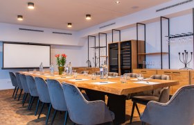 Moderner Konferenzraum mit großem Holztisch, grauen Stühlen und Präsentationsleinwand.